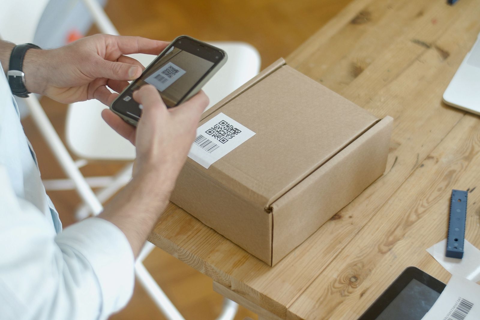 Mano con reloj pulsera fotografiando el código QR de una caja con el celular para registrar el stock